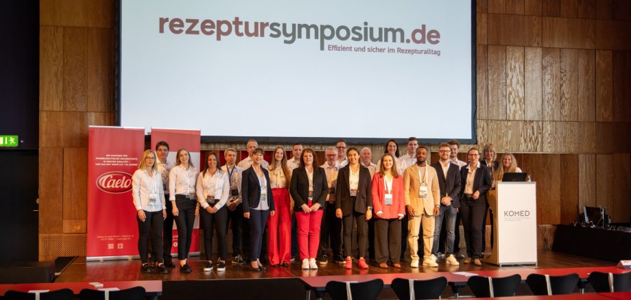 Eine große Gruppe von Mitarbeitern von Caelo und WEPA, die auf einer Bühne stehen. Dahinter eine Leinwand, auf der der Schriftzug rezeptursymposium.de steht.