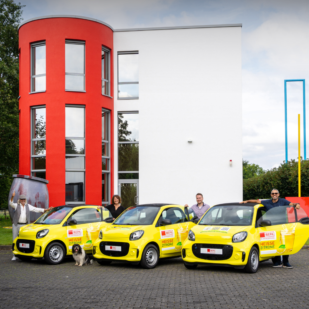 WEPA Smart Gewinnspie Übergabe - 2 Drei gelbe Smart-Autos stehen nebeneinander, jedes mit einem Schriftzug. Neben jedem Auto stehen Personen, und vor einem der Fahrzeuge sitzt ein Hund.