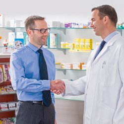 Zwei Männer schütteln sich die Hand. Ein Man trägt eine Brille und ein blaues Hemd und der andere Mann ist gekleidet wie ein Apotheker in langen weißen Kittel.