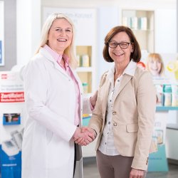 Eine Apothekerin und eine Kundin schütteln sich die Hand und schauen lachend in die Kamera