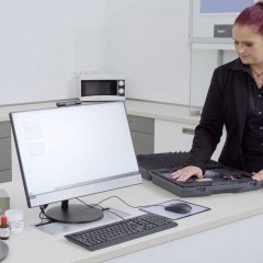 Eine Frau mit schwarzen Anzug baut ein Labor auf. Ein Bildschirm mit Tastatur steht auf der Anrichte.