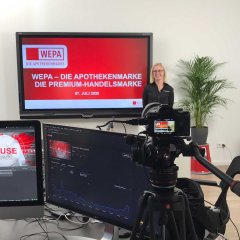 Eine blonde Mitarbeiterin der WEPA steht mit schwarzer WEPA Bluse mit roten Logo an den Hals gestickt vor einem Bildschirm. Sie präsentiert ein Online-Event. Auf Sie wird eine Kamera gerichtet.