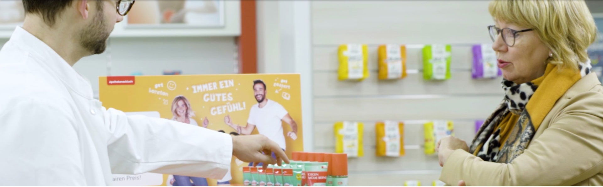 Ein Apotheker und eine Kundin stehen vor einem WEPA-Die Apothekenmarke Pferdedisplay. Der Apotheker zeigt der Kundin die Pferdesalbe von WEPA.