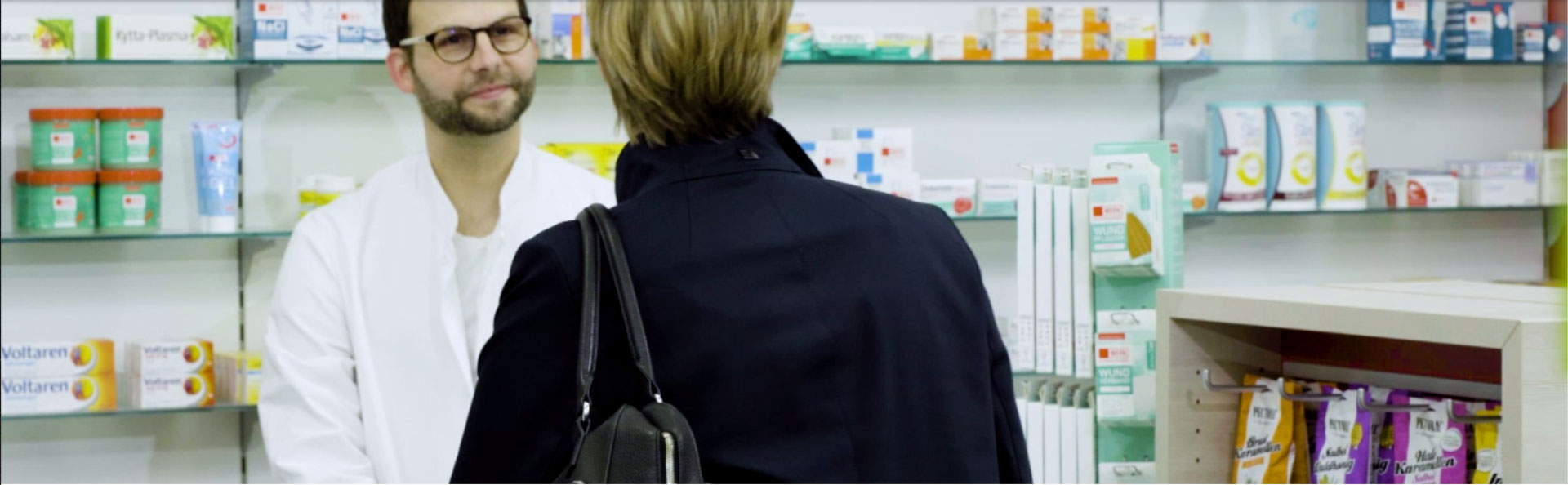 Über uns Eine Kundin steht vor einem Apotheker am HV Tisch und wird beraten