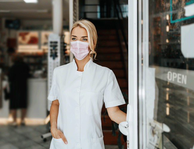 Apothekerin mit Mundschutz und Kittel öffnet eine Apothekentür.