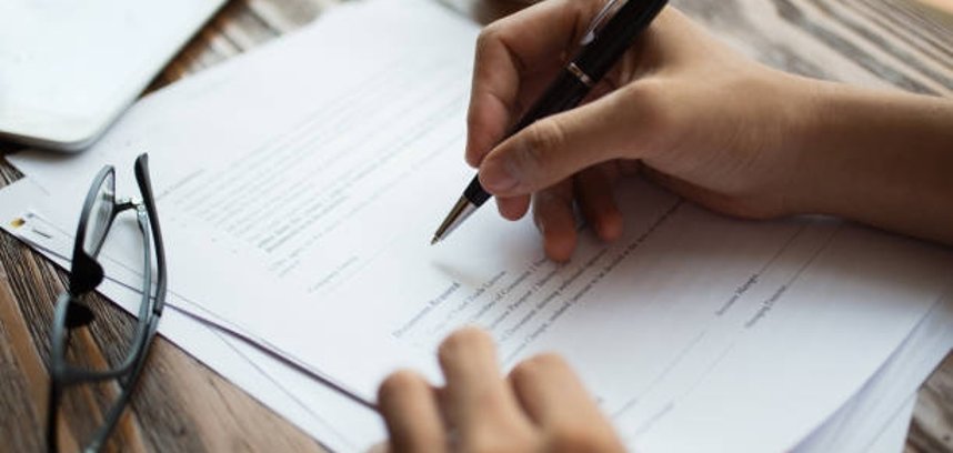 Unrecognizable businessman examining papers at table. Manager with ballpoint pen filling business papers. Close-up of male hands working at desk. Analyzing documents concept