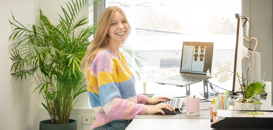 Eine Person steht an einem Computer und lächelt in die Kamera.