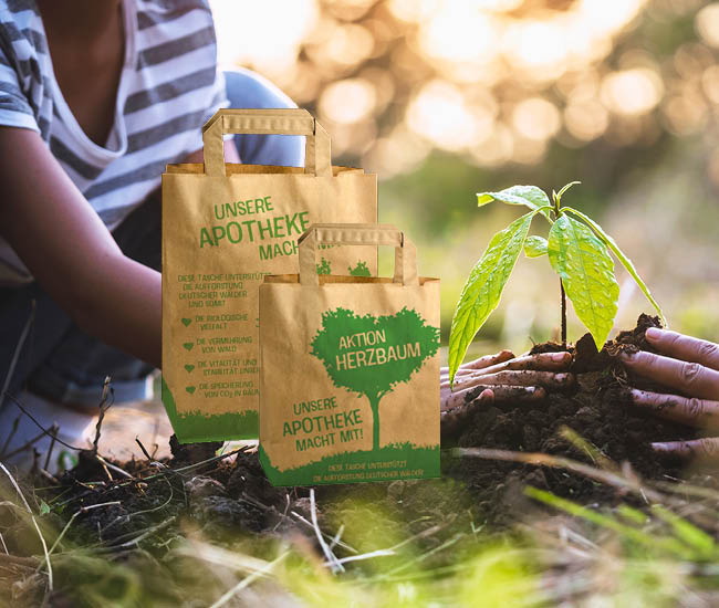2 Tragetaschen von Aktion Herzbaum vor einer Hand die eine Pflanze in die Erde setzt